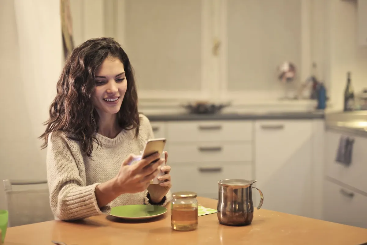 Uśmiechnięta kobieta przy kuchennym stole patrzy w telefon, obok herbata